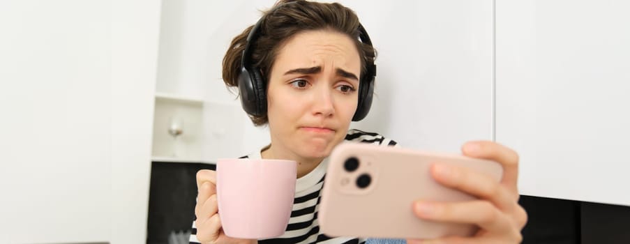 Close up portrait of young woman in headphones, watching something sad on smartphone application, frowning and looking with sympathy at mobile phone screen, drinking tea or coffee in the kitchen.