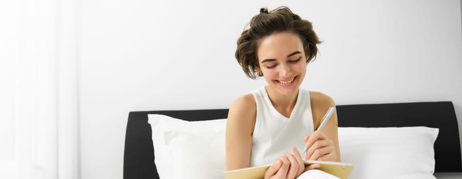 Portrait of smiling, happy young woman sitting in bed, writing in journal, making notes in diary, doing homework in bedroom.
