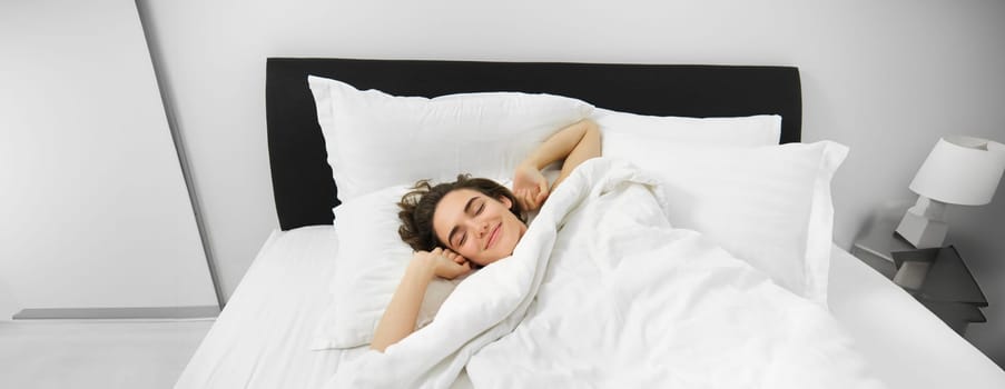 Portrait of smiling, pleased young woman stretching her arms, waking up after good night sleep, lying on soft white pillow in bed with comfortable linen sheets.