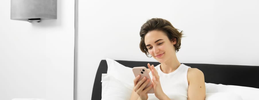 Portrait of stylish, modern young woman at home, sitting on bed and using mobile phone, reading on smartphone, scrolling social media application in bedroom.