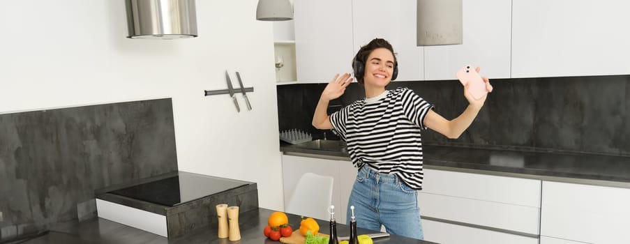 Happy woman dancing and cooking in the kitchen, listening music in earphones, making a meal, preparing salad, chopping vegetables on a counter.