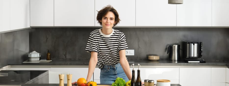 Portrait of beautiful young woman, female model posing near chopping board with vegetables and salad dressing, making herself light diet meal, vegan food for breakfast, standing in kitchen.