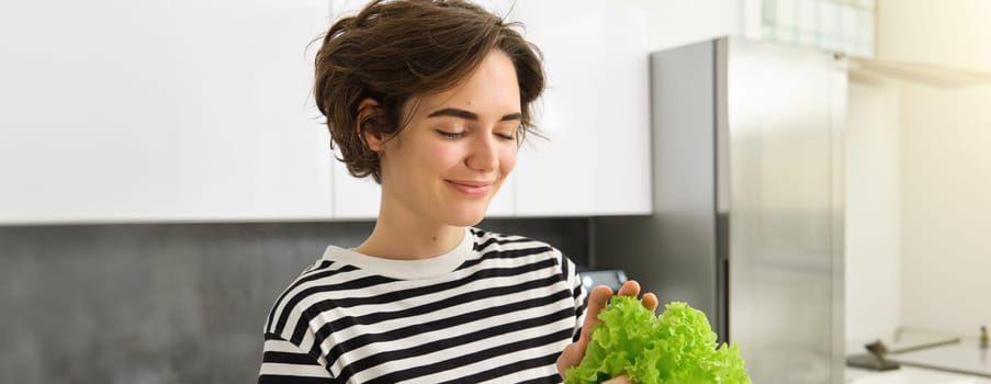 Portrait of young smiling woman in the kitchen, holding fresh green leaves of lettuce, preparing vegetables for salad, cooking healthy vegetarian meal.