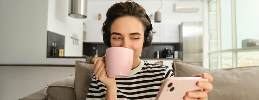 Close up of woman watching video with interest, looking at smartphone screen, wearing headphones and drinking tea from pink cup, sitting on sofa in living room at home.