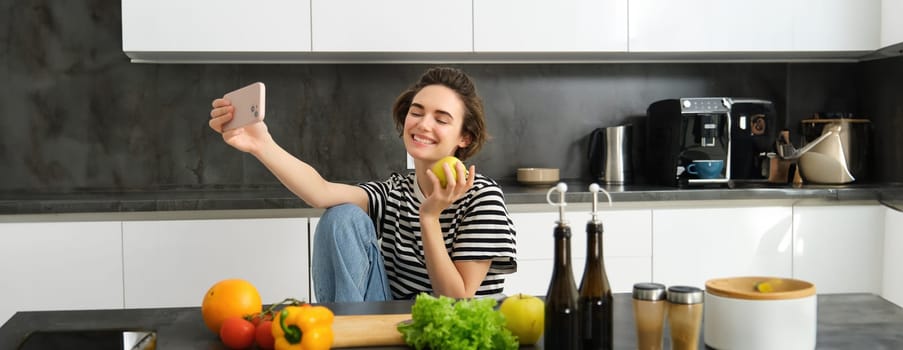 Cute young woman, modern lifestyle blogger, taking selfie while cooking salad in the kitchen, using smartphone to make photos for social media app.