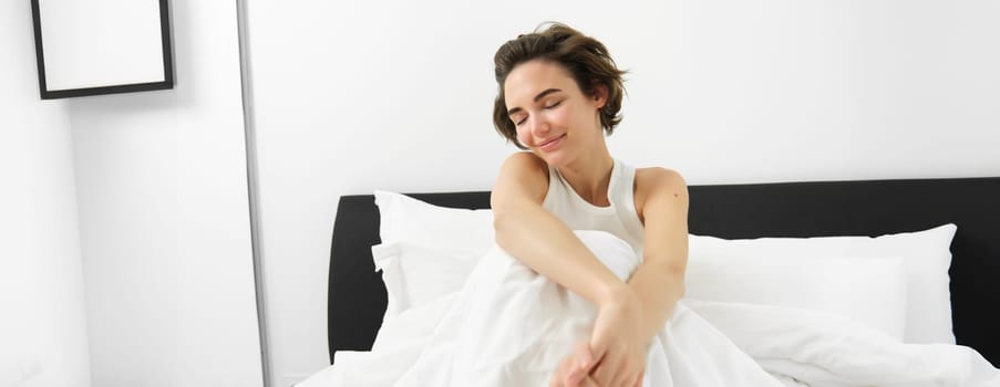 Portrait of cute smiling woman, sitting in bed, resting at home, lying under white linen sheets, waking up in morning with happy, pleased face expression.