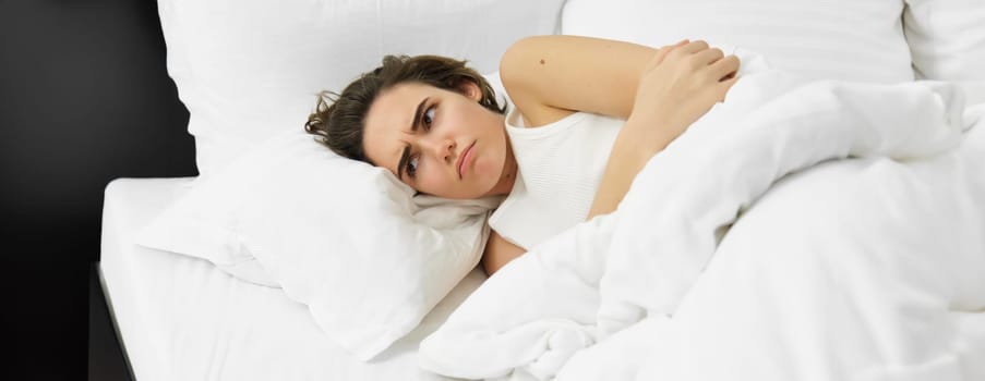 Close up of upset woman lying in bed with menstrual cramps, has period pain, touching her stomach and frowning, covered in white duvet.