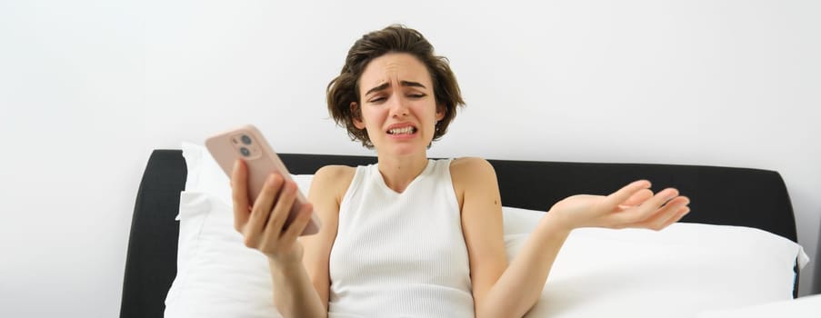 Portrait of woman with confused face, laying in bed with smartphone, chatting on mobile phone app, video call, shrugging shoulders at some nonsense, feeling upset by something she sees online.