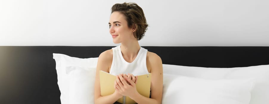 Portrait of young sensual woman, hugging her notebook, writing in journal or diary, smiling and looking aside, sitting on bed, noting her dreams or feelings in morning.