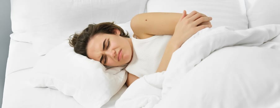 Young woman lying in bed with stomach ache, has painful cramps, menstrual pain. Lifestyle and wellbeing concept
