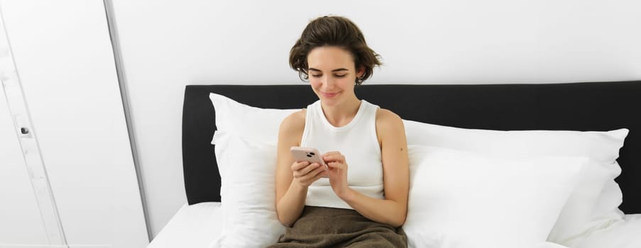 Portrait of young beautiful brunette woman, sitting on bed with smartphone, using mobile phone app, sending message and smiling, reading on telephone application in bedroom.