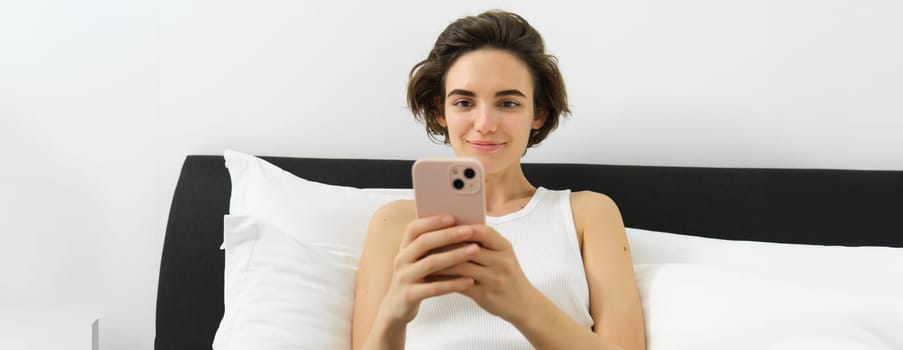 Portrait of smiling brunette woman resting in her bedroom, using mobile phone and lying in bed in her home outfit, messaging, sending message on smartphone app.