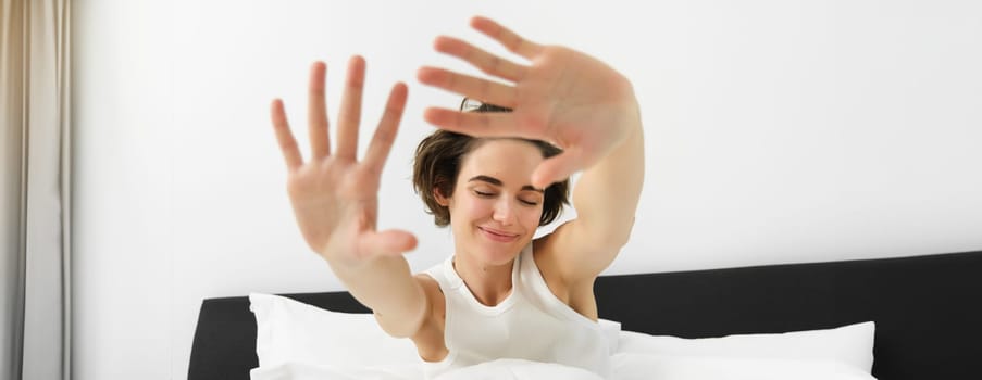 Close up portrait of carefree, beautiful young woman stretching her arms and smiling with dreamy, closed eyes, waking up in morning after good night sleep.