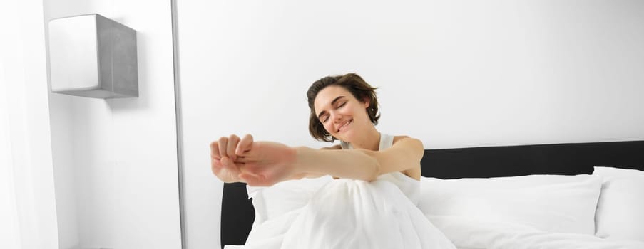 Portrait of smiling young woman wakes up in her bed, sitting under white linen sheets with closed eyes and stretching her hands with satisfaction.