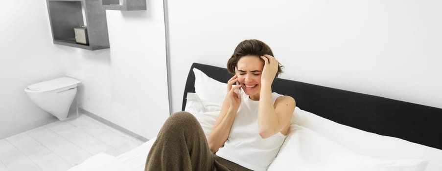 Beautiful young modern woman talking to friend, lying in bed and calling someone on phone, resting at home, chatting.