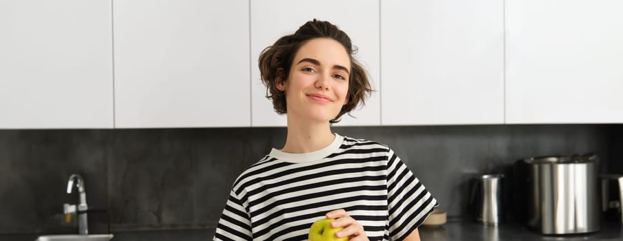 Portrait of beautiful, smiling young woman, holding an apple, eating fruit in the kitchen, looking happy. Concept of healthy diet and lifestyle.