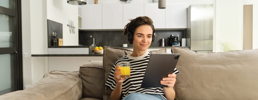 Portrait of woman in headphones, sitting on sofa with tablet and drinking orange juice, watching tv show on her gadget, using social media application, spending time at home.
