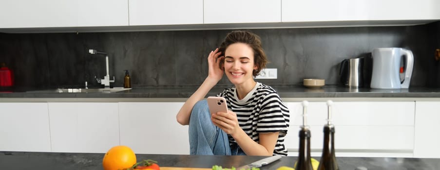 Portrait of young woman smiling, laughing and using smartphone while cooking, searching recipes on smartphone app, preparing meal from vegetables in her kitchen at home.