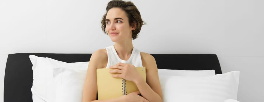 Image of mindful young woman sitting in bed, waking up and writing in journal, making notes, sharing her thoughts in diary.