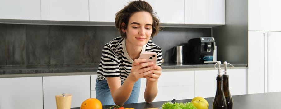 Portrait of young smiling woman, searching cooking recipe online on smartphone, standing near vegetables and chopping board, making meal, preparing salad and using social media on mobile phone.