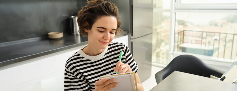 Close up portrait of smiling, cute woman with notebook, writing down recipe, making notes or grocery list for shopping, sitting in the kitchen.