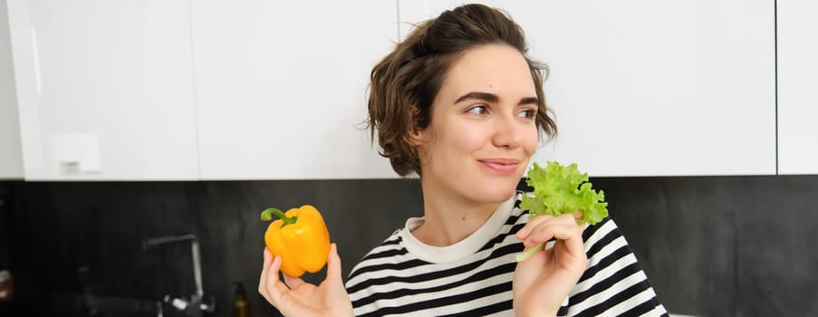 Portrait of smiling brunette woman, holding lettuce leaf and yellow sweet pepper, eating vegetables, likes fresh vegies, healthy diet concept.