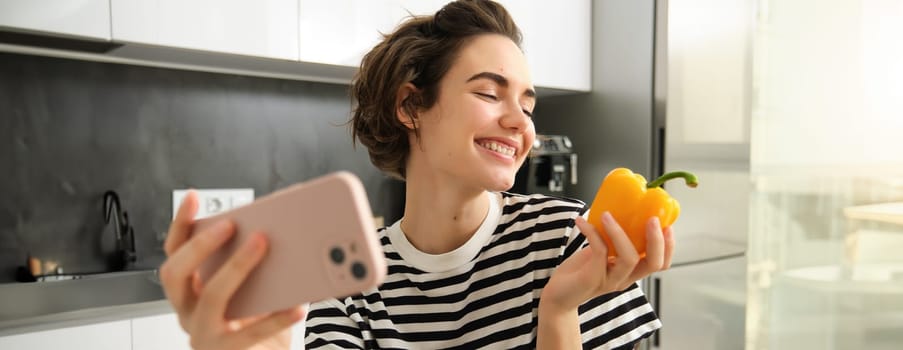 Smiling modern woman, vegetarian food blogger, taking selfie with fresh yellow sweet pepper, holding smartphone, recording a video blog for her social media account, posing in the kitchen.