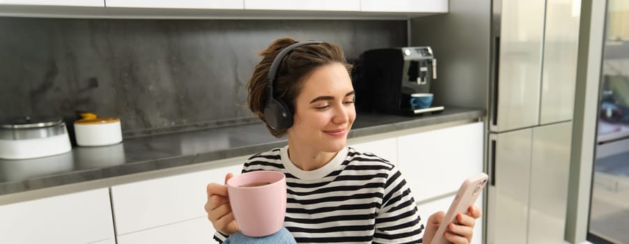 Close up portrait of young female student, drinking tea and listening to music in headphones, using smartphone in kitchen.