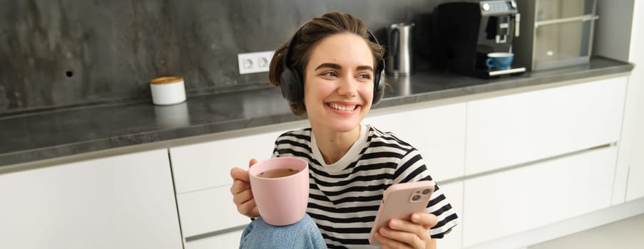 Stylish young woman in headphones, listens music and uses smartphone, drinks warm tea in kitchen, relaxing at home.