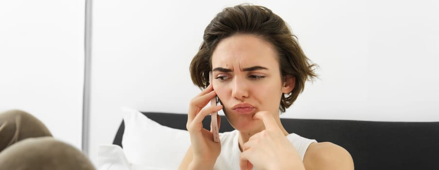 Close up portrait, woman makes a phone call with puzzled, frowning face, hearing complicated story over the telephone, talking to someone while resting in bed.