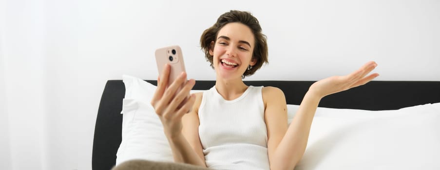 Smiling friendly young woman chatting on smartphone app, talking via video call app on mobile phone, lying in bedroom, resting in bed and communicating with someone.