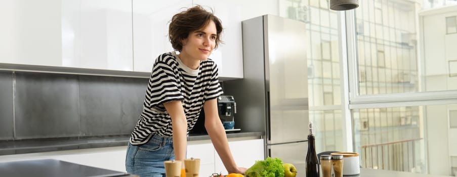 Portrait of beautiful young woman, female model posing near chopping board with vegetables and salad dressing, making herself light diet meal, vegan food for breakfast, standing in kitchen.