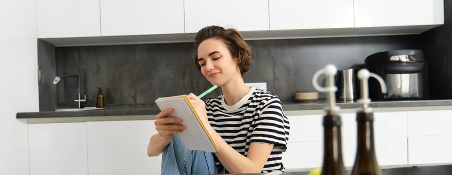 Portrait of beautiful modern woman, girl cooking with vegetables, holding notebook, writing down recipe, making notes, dinner plans, creating meal list for weekend, sitting in kitchen.