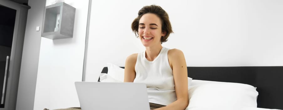 Modern woman resting at home, sitting on bed with laptop, looking at computer screen, laughing and smiling.