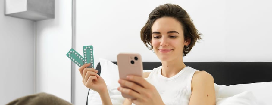 Smiling female model, holding pills medication and looking at her phone, reading prescription on mobile app, resting in bed.