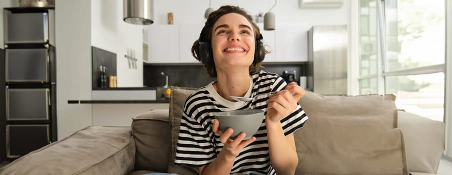 Close up of happy laughing woman, smiling, eating in front of tv, holding bowl and spoon, wearing wireless headphones, watching favourite tv show on big screen in living room.