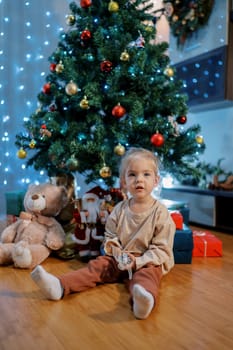 Little girl with a toy in her hand sits on the floor near the Christmas tree. High quality photo