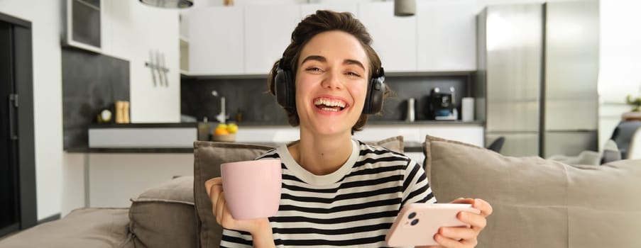 Close up portrait of happy woman with smartphone and headphones, drinking tea and watching videos on social media app, laughing and smiling.