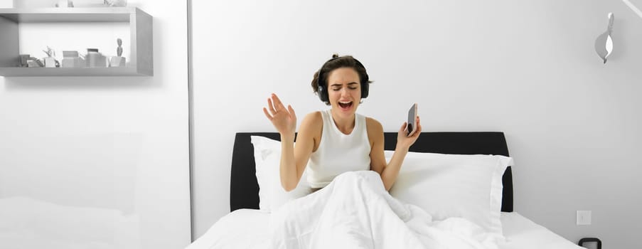 Portrait of happy woman singing in her bed, wakes up and listens to music in wireless headphones, holding mobile phone.