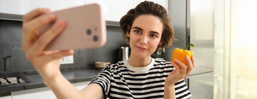 Close up portrait of cute brunette woman, taking selfie with yellow fresh pepper, recording video on smartphone, posing with vegetables in the kitchen, making cooking blog on social media app.