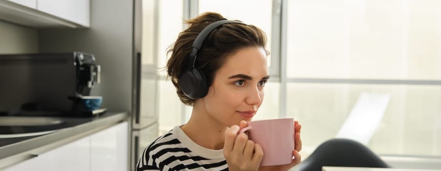 Close up portrait of smiling brunette woman, student drinks her tea and listens music or ebook in headphones, sits in kitchen and relaxes.