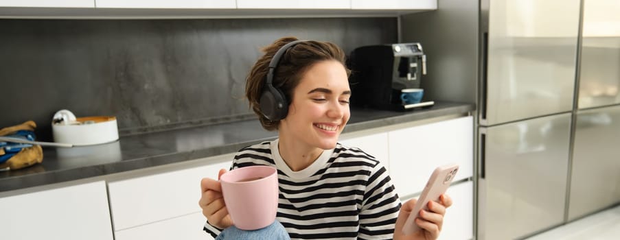 Portrait of smiling young woman, using mobile phone, sitting in kitchen and drinking hot tea, browsing social media and relaxing at home.