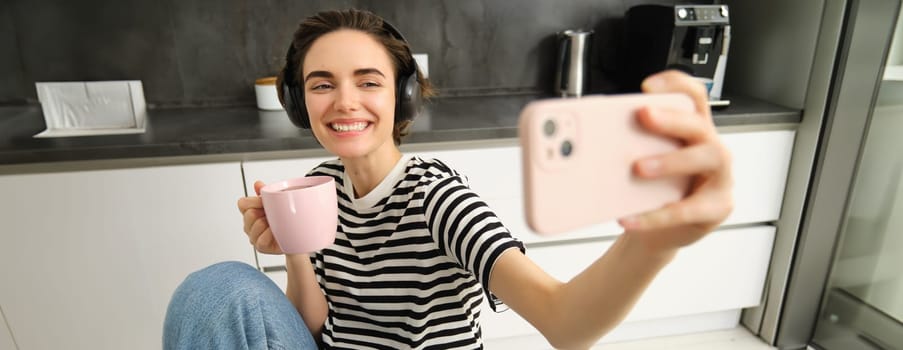 Portrait of modern happy woman, taking selfie, lifestyle blogger recording video of her eating breakfast and drinking tea in the kitchen, using smartphone for making content.