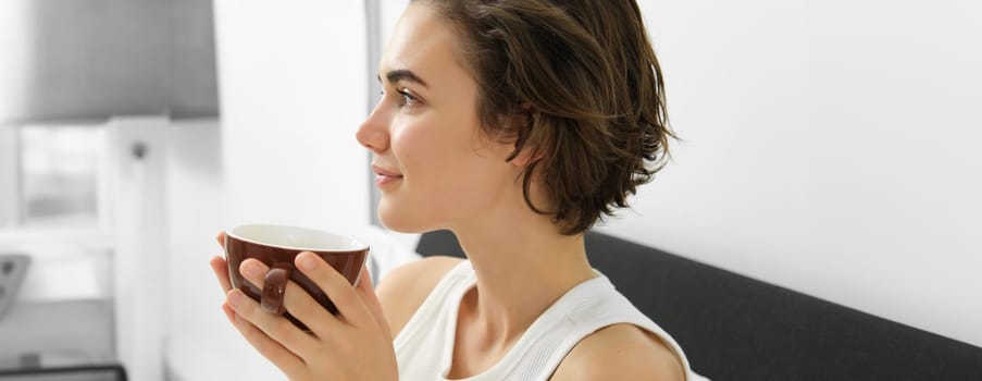 Sensual morning concept. Young beautiful woman drinking coffee in bed, holding mug and smelling nice aroma of freshly brewed drink.