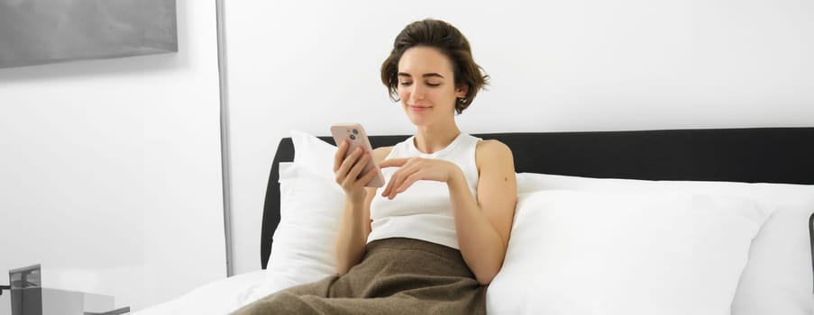 Portrait of cute female model in bedroom, holding phone, typing message on smartphone, resting in bed and smiling.