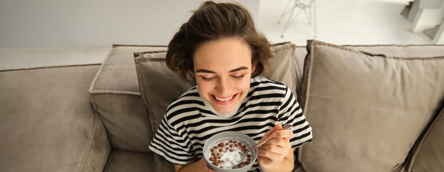 Close up of cute young female model, eating cereals with milk, enjoys her breakfast on sofa in living room, smiling and looking happy. People and food concept