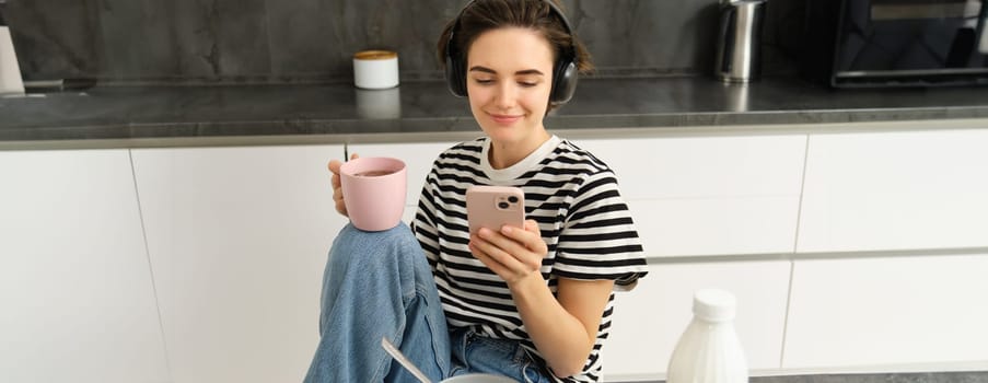 Portrait of young woman drinking tea and listening to music in headphones, scrolling social media while having lunch in kitchen, smiling happily.