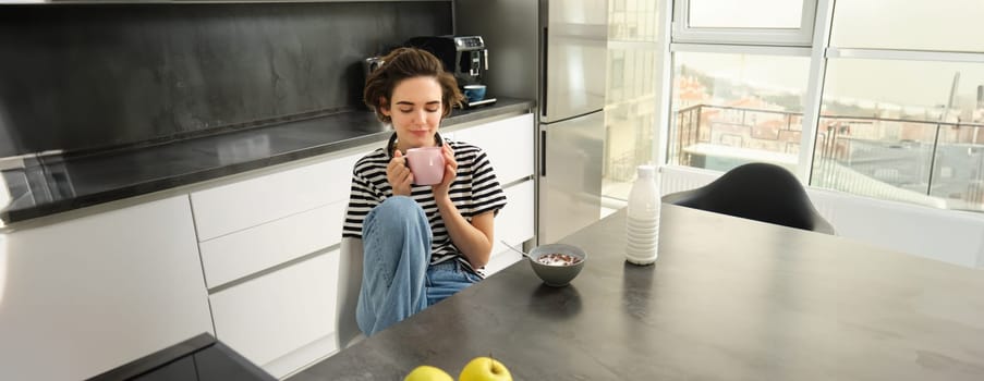 Cute brunette woman, student eating breakfast in morning, drinking hot tea, smiling and looking pleased.