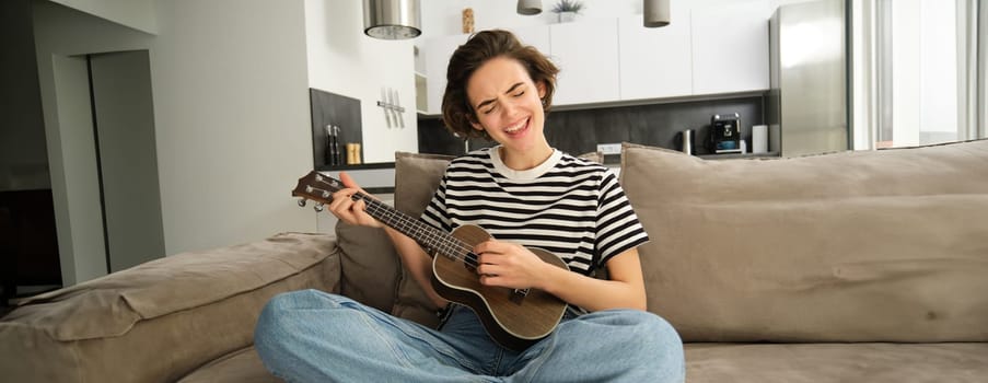Portrait of cheerful young woman playing her ukulele, singing and laughing, sitting in living room at home. Lifestyle and music concept