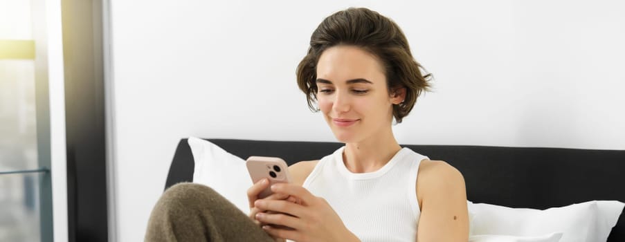 Portrait of young beautiful brunette woman, sitting on bed with smartphone, using mobile phone app, sending message and smiling, reading on telephone application in bedroom.